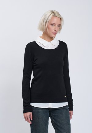 Kvinde med kort blondt hår iført en hvid skjorte med krave under en sort sweater og mørke jeans, stående mod en ensfarvet grå baggrund.