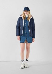 Giacca puffer blu marina con fodera rosa chiaro, vestito di denim con bottoni, calze bianche con strisce nere e scarpe da ginnastica chiare.