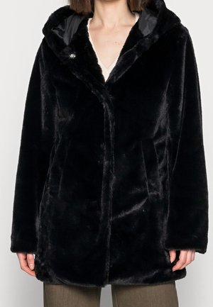 Žena oblečená v černém kabátě s umělým kožíškem a kapucí přes béžový top a hnědé kalhoty, ruce viditelné po stranách.