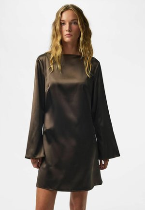 Femme aux longs cheveux blonds ondulés, portant une robe ample en satin marron foncé brillant à manches longues, sur un fond clair uni.