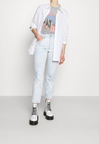 Persona vestida con vaqueros de mezclilla azul claro, botas blancas de cordones, camiseta gráfica gris y una camisa blanca de botones con las mangas arremangadas, de pie con una mano en el bolsillo.