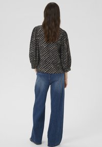 Hosszú barna hajú nő fekete mintás blúzban, puffos ujjakkal és bő szárú kék farmerban, egy sima háttér előtt áll.