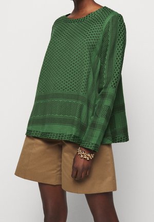 Osoba nosząca zielony, długi rękaw, teksturowany top z wzorzystymi detalami, beżowe krótkie spodenki do kolan oraz złotą bransoletkę z koralików na lewym nadgarstku.