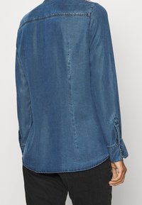 Jeansskjorta i mörkblått med en slät textur, knäppta manschetter och en rundad nederkant. Har en klassisk krage och diskreta stickningsdetaljer.