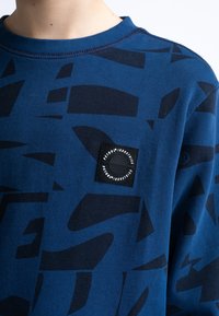 Felpa blu con motivo astratto nero, colletto a costine, e un'etichetta quadrata con un logo circolare sul petto. Tessuto morbido.