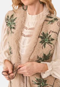 Chaleco beige con patrones florales verdes bordados y lentejuelas, con un suave borde de piel. Blusa blanca debajo con detalles de encaje.