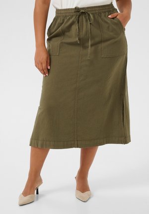 Mujer que lleva una falda midi verde oliva con bolsillos delanteros, cintura con cordón, abertura lateral y zapatos de tacón beige, de pie sobre un fondo liso.