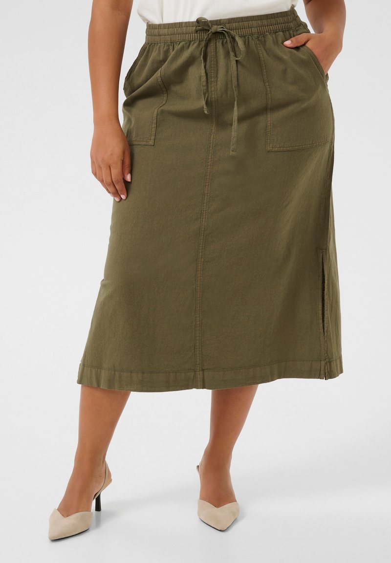 Femme portant une jupe midi vert olive avec poches avant, taille coulissée, fente latérale et chaussures à talons beiges, debout sur un fond uni.