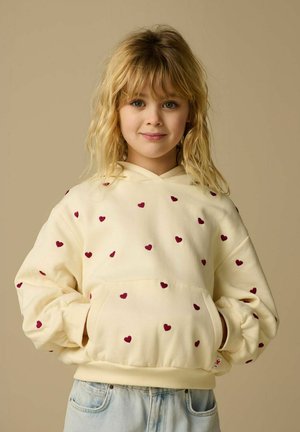 Jeune fille aux cheveux blonds ondulés portant un sweat à capuche crème avec des motifs de cœurs rouges et un jean bleu clair, debout devant un fond beige.