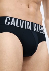 Čierne bavlnené slipy so širokým šedým logom Calvin Klein. Hladká textúra, priliehavý strih a predný zásobník pre podporu.