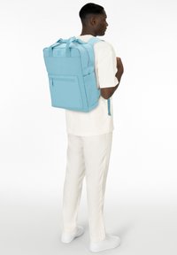 Zaino rettangolare blu chiaro con spallacci regolabili, tasca frontale con zip e tessuto testurizzato. Indossato da una persona in un outfit bianco.