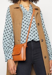 Un gilet sans manches marron sur un blouse à motifs floraux bleu clair. Un jeans en denim foncé. Un petit sac bandoulière orange avec des accents texturés.
