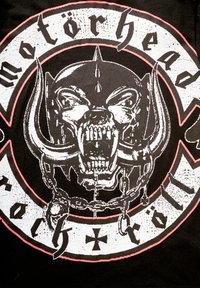 Juodas marškinėliai su grafiką, kurioje pavaizduota �įnirtinga kaukolė su iltimis, grandinėmis ir tekstu „motörhead“ paryškintu rašmenimis, apibrėžtu raudona spalva.