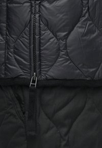 Chaqueta acolchada negra con cuello alto, cierre de cremallera y patrones de costura en contraste. La tela parece suave y resistente al agua.