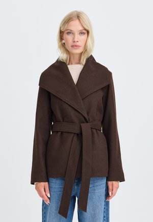 Femme blonde portant une veste marron foncé cintrée avec un large col, sur un haut beige et un jean bleu, sur un fond clair uni.