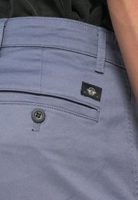 Pantalones de tela gris claro con diseño de frente plano, cierre de botón y una etiqueta negra con un logo en el bolsillo trasero.
