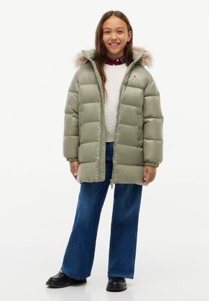 Ragazza sorridente con giacca imbottita verde con cappuccio di pelliccia, maglione bianco, jeans blu e scarpe nere, in piedi davanti a uno sfondo bianco.
