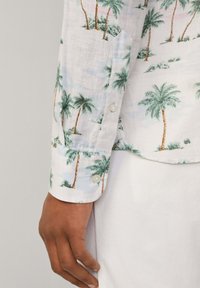 Camisa de lino blanca con un patrón de palmeras, acentos en verde y marrón. Puño abotonado con botones de color claro, textura suave.