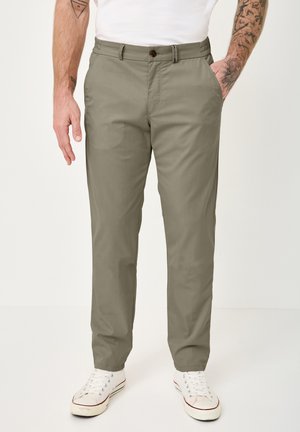 Man wearing light olive chinos and white sneakers, standing with one hand in pocket and tattoos visible on forearms.