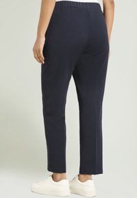 Person in schwarzen geraden Hosen und weißen Sneakers, von hinten vor einem schlichten Hintergrund gezeigt.