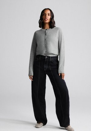 Woman standing with hands by sides wearing a gray buttoned cropped sweater, wide-leg black jeans, and white patterned shoes on plain background.