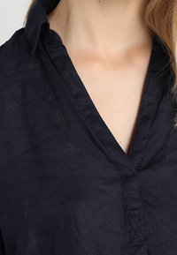 Marineblauwe linnen blouse met een V-hals, geplooide kraag en subtiele stikdetails. De stof heeft een licht gestructureerde afwerking.