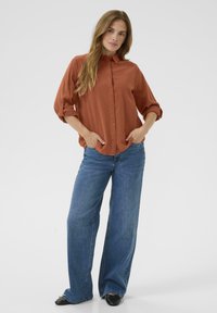 Roestkleurig, knoopshirt met een kraag en opgerolde mouwen, gecombineerd met wijde blauwe jeans en zwarte schoenen. Eenvoudig ontwerp, casual pasvorm.
