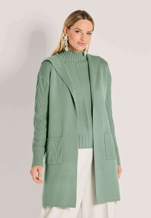 Vrouw in een lichtgroene gebreide cardi met zakken over een bijpassende hoge trui en witte broek, poserend tegen een effen achtergrond.