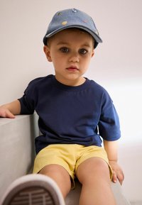 Mörkblå bomullströja i kombination med gula shorts. Barnet har på sig en denimkeps med färgglad broderi. Sitter mot en ljus bakgrund.