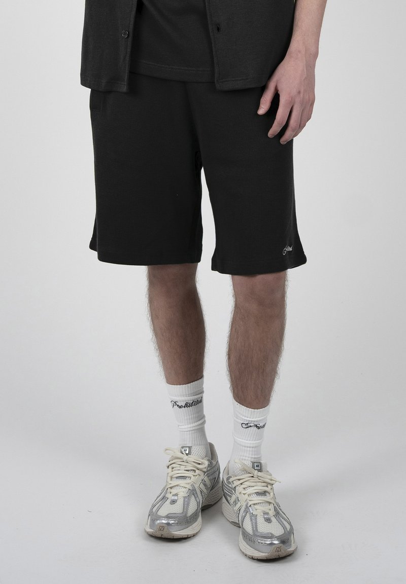 Homme portant un short noir, des chaussettes blanches avec texte, et des baskets New Balance argentées-grises, debout sur un fond clair uni.