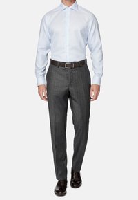 Uomo con camicia a maniche lunghe azzurra chiara, pantaloni grigi a righe sottili, cintura marrone scuro e scarpe da uomo in pelle marrone scuro, in piedi davanti a uno sfondo bianco.