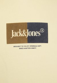 T-shirt di colore crema con un logo rettangolare con strisce verticali marroni e blu navy. Il testo include "Jack & Jones" e altri dettagli di branding.