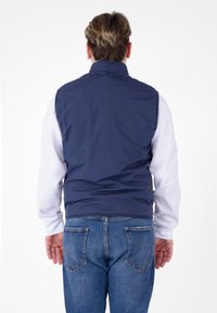 Gilet senza maniche di colore blu navy realizzato in tessuto liscio, con colletto alto e orlo che arriva alla vita, indossato sopra una camicia bianca a maniche lunghe.