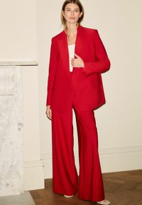 Sacou roșu adaptat și pantaloni largi, dintr-un material texturat. Întreaga ținută are un design structurat, cu buzunare și o siluetă ajustată.
