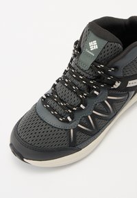 Scarpa da ginnastica da uomo con tomaia in mesh grigia, dettagli neri, lacci rotondi e suola imbottita con finitura testurizzata.