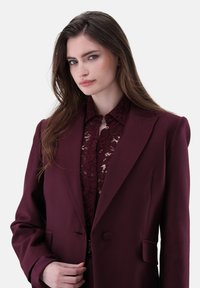 Blazer bordeaux con vestibilità su misura, rever a punta e bottoni frontali. Sotto, una camicia in pizzo bordeaux con dettagli di bottoni.