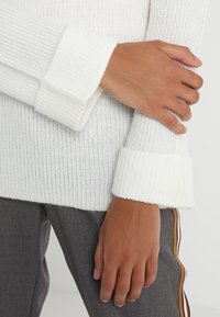 Valkoinen urheiluvalkoiset neulepaita, jonka leveät, käännetyt hihansuut korostavat pehmeää tekstuuria ja rentoa istuvuutta, yhdistettynä harmaisiin housuihin, joissa on sivuvaihtoehtoja.