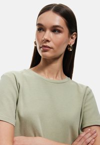 T-shirt in cotone verde chiaro con maniche corte, caratterizzato da un colletto rotondo e una texture liscia. Il modello indossa orecchini a cerchio dorati.
