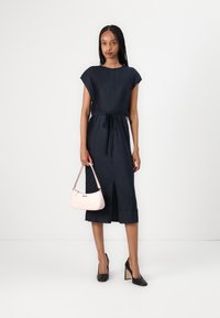 Abito a coste navy con maniche a cappuccio, cintura in vita e spacco frontale; abbinato a una borsa rosa chiaro e tacchi neri.