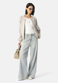 Chaqueta bomber gris claro sobre una camiseta blanca, combinada con unos vaqueros de pierna ancha de color azul claro y zapatos de punta blanca. Sosteniendo un pequeño bolso beige.