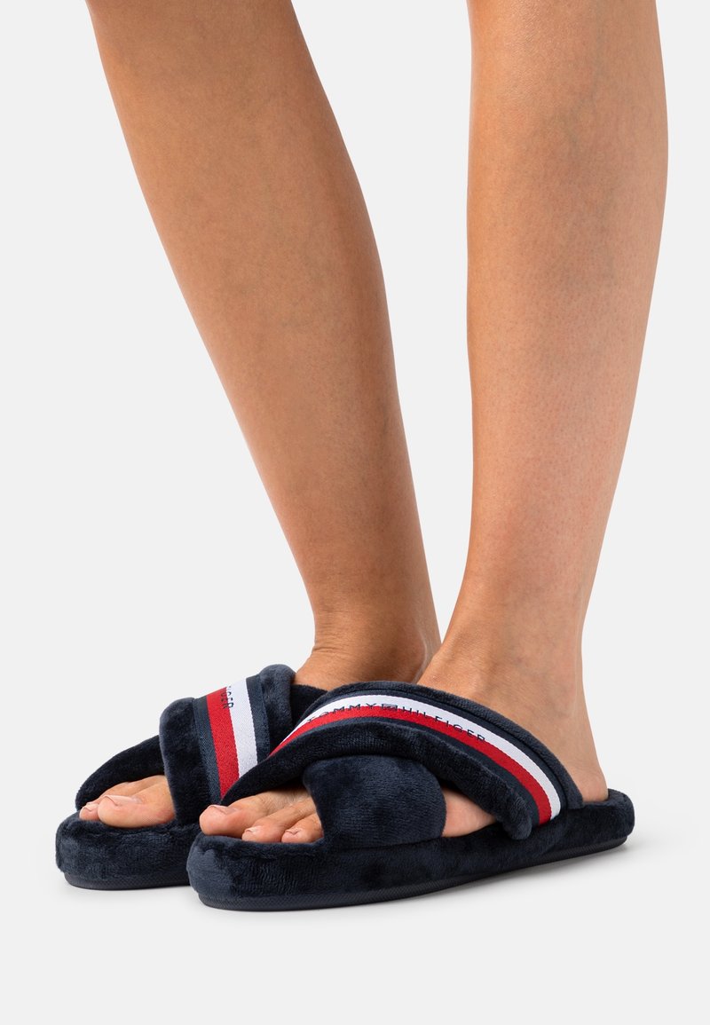 Pieds nus portant des chaussons en peluche bleu marine avec des brides rayées rouge, blanc et bleu et le logo Tommy Hilfiger sur un fond uni.