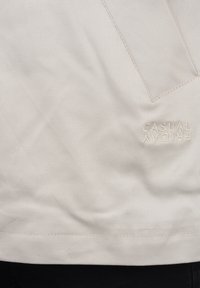 Lys beige stoff med en subtil tekstur og en liten, brodert logo som sier "CASUAL FRIDAY" nær kanten.