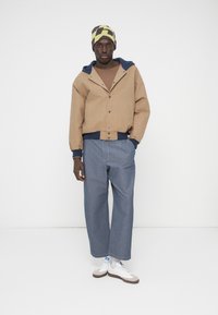 Hnedá skrátená bunda s námorníckymi modrými detailmi, kombinovaná so svetlomodrými širokými rifľovými nohavicami a bielymi teniskami. Vzorovaná čiapka v žltej a hnedej farbe.