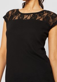Kvinna som bär en svart kortärmad topp med blommig spetsdetalj över övre bröstet och axlarna.