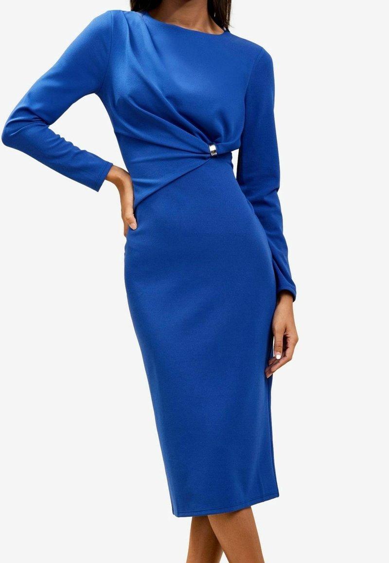 Femme portant une robe midi ajustée à manches longues bleu roi avec un pli froncé et un détail boucle argentée à la taille.