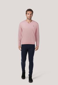 Lichtroze V-hals trui met lange mouwen, slim fit ontwerp, gedragen over donkere blauwe slim-fit jeans en zwarte laarzen. Subtiel logo-detail op de borst.