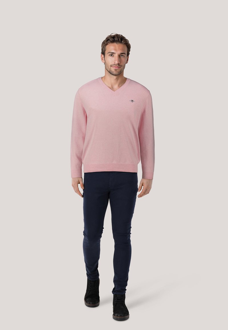 Lichtroze V-hals trui met lange mouwen, slim fit ontwerp, gedragen over donkere blauwe slim-fit jeans en zwarte laarzen. Subtiel logo-detail op de borst.
