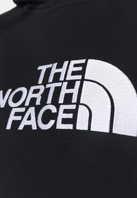 Černá mikina s bílým vyšívaným logem „THE NORTH FACE“ a designem polokruhu. Hladká struktura, pohled zblízka.