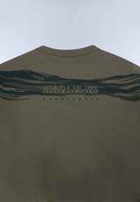 Camiseta verde oliva de manga corta, con un estampado oscuro ondulado y el texto "NAPAPIURI geographic" en la parte posterior. Material de algodón, diseño casual.