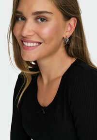 Zwarte ribbel top met een ronde halslijn, verfraaid met een klein zwart strikje. Model heeft lang bruin haar en zilveren creolen.
